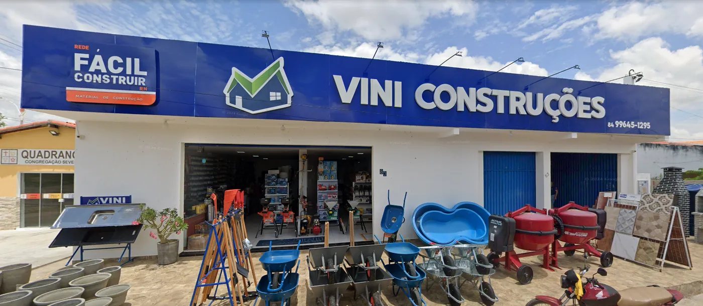 Fachada azul da Vini Construções em Severiano Melo, RN, com carrinhos de mão, betoneiras e pisos expostos na calçada
